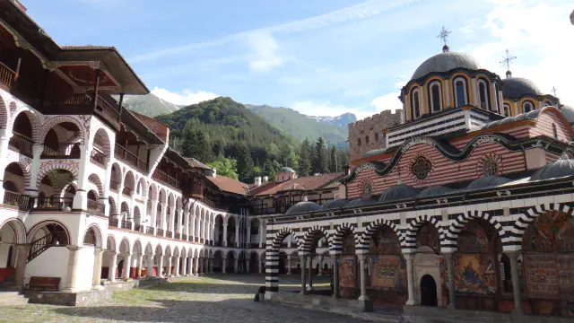 Courtyard of Rila Monastery with arcaded wooden galleries on the left, the striped arches and domes of the main church on the right and forested mountains behind