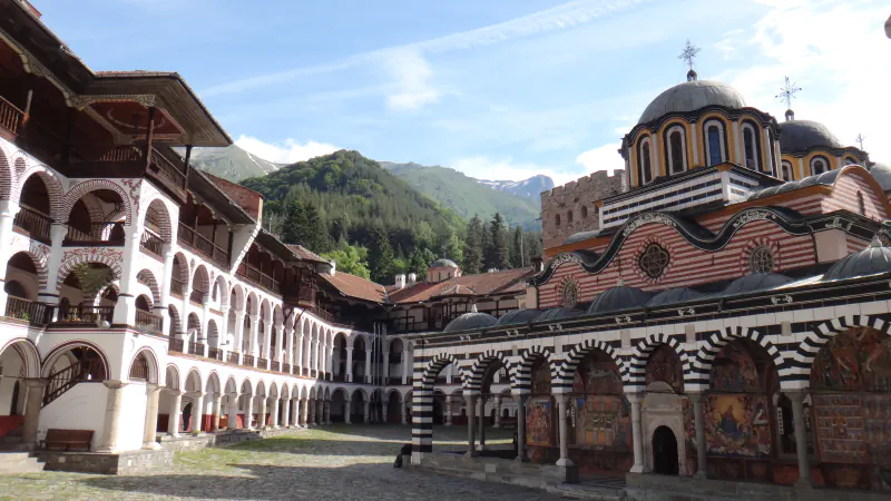 Courtyard of Rila Monastery with arcaded wooden galleries on the left, the striped arches and domes of the main church on the right and forested mountains behind