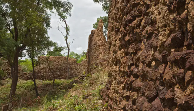 Rough dark-brown laterite stone wall filling the right of the frame, with a lower stone wall, a few thin trees and grass beyond it under a cloudy sky