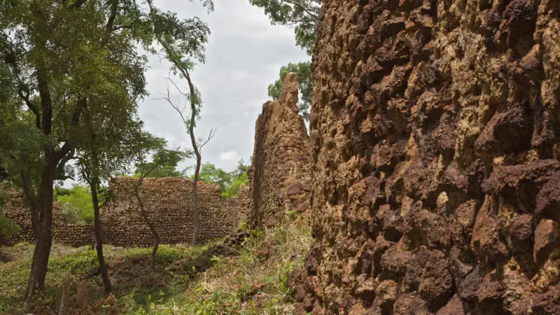 Rough dark-brown laterite stone wall filling the right of the frame, with a lower stone wall, a few thin trees and grass beyond it under a cloudy sky