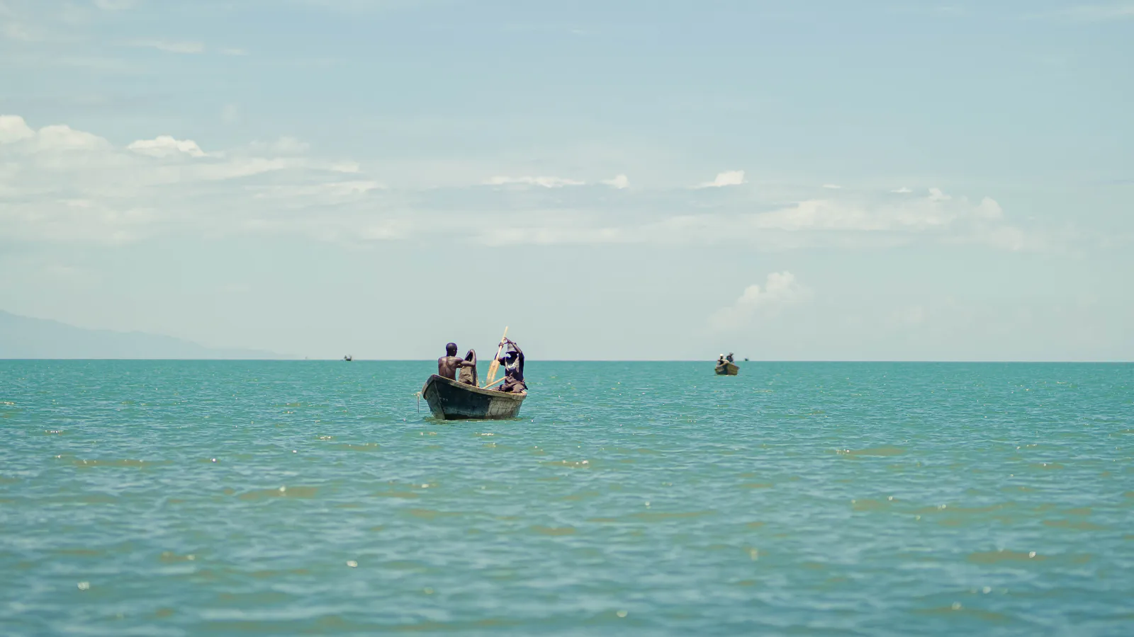 A wooden fishing canoe with two people on calm turquoise water, a second canoe farther out, a hazy shoreline at the left edge and a pale sky with thin clouds