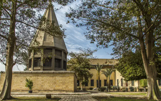 A pale stone base with an octagonal timber pavilion and a tall faceted pointed roof stands in a garden among trees; a low yellow building is behind it