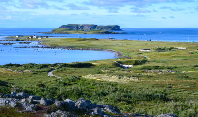 Grassy coast with a curving bay, boardwalk paths and a fenced area on the right, with a low flat-topped island offshore under a blue sky and rocks in the foreground