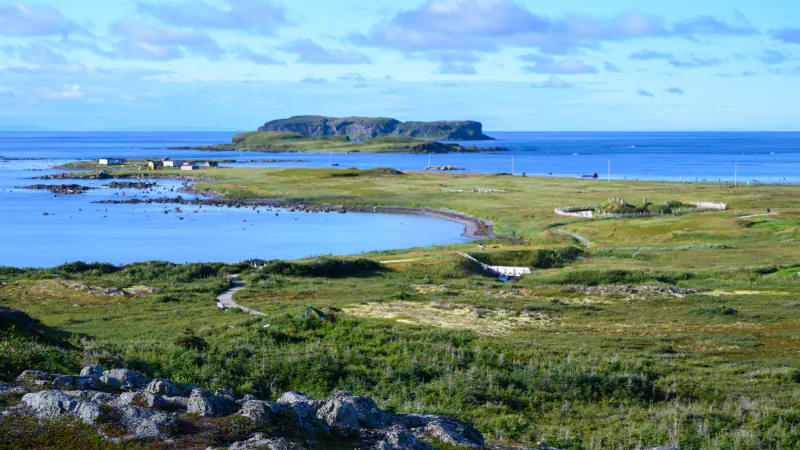 Grassy coast with a curving bay, boardwalk paths and a fenced area on the right, with a low flat-topped island offshore under a blue sky and rocks in the foreground