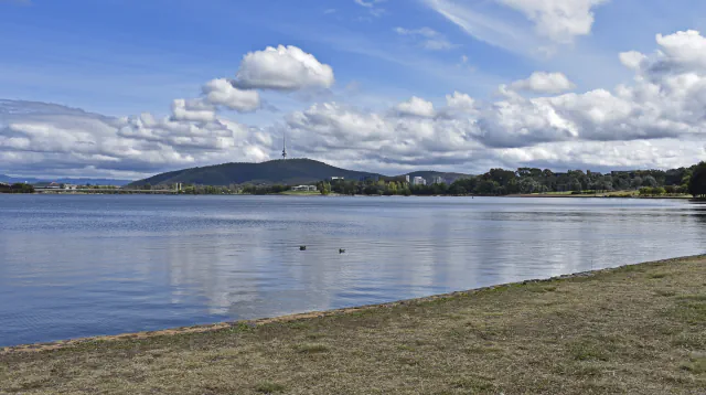 Lake Burley Griffin, grassy shore and a hill with a tower under a cloudy sky