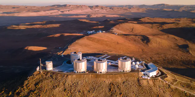 Aerial view at sunset of the four large Unit Telescope enclosures of the Very Large Telescope on Cerro Paranal, with brown desert hills reaching to the horizon