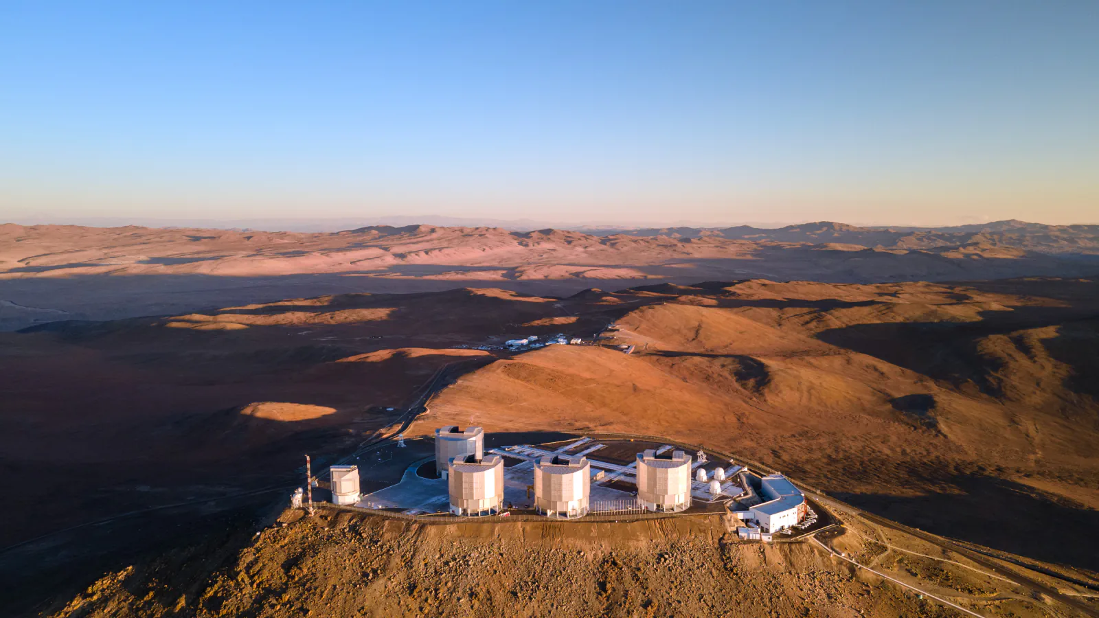 Pogled iz zraka ob sončnem zahodu na štiri velike stavbe teleskopov Very Large Telescope na gori Cerro Paranal; rjavi puščavski griči segajo do obzorja