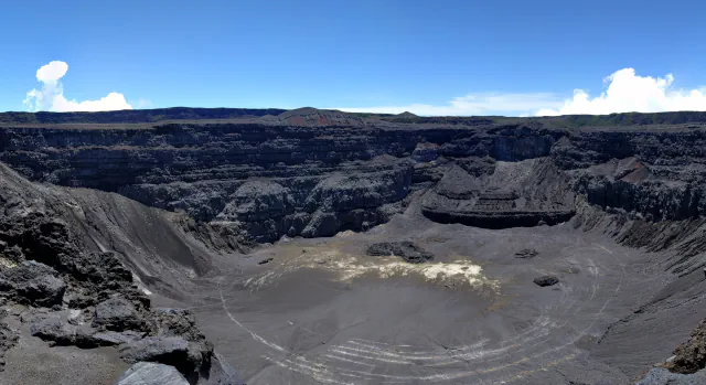 A wide panorama into a deep volcanic crater, with dark layered walls, a grey crater floor marked by pale patches and curved ridges, beneath a blue sky with a few clouds