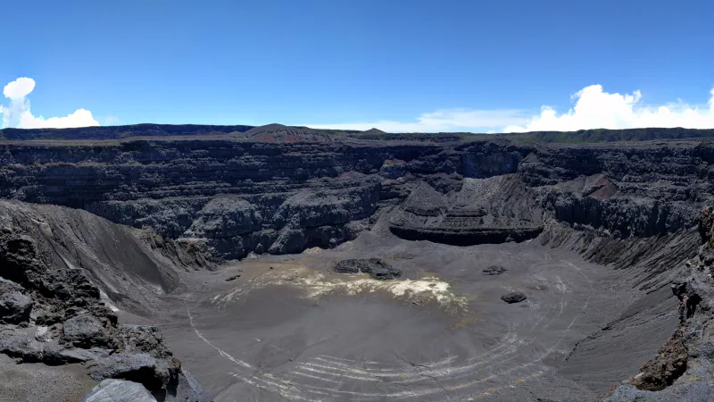 A wide panorama into a deep volcanic crater, with dark layered walls, a grey crater floor marked by pale patches and curved ridges, beneath a blue sky with a few clouds