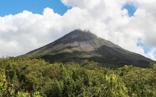Arenal volcano’s steep, dark cone rises above green forest, with the summit partly hidden by cloud
