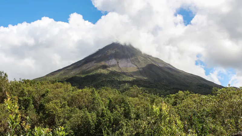 Arenal volcano’s steep, dark cone rises above green forest, with the summit partly hidden by cloud