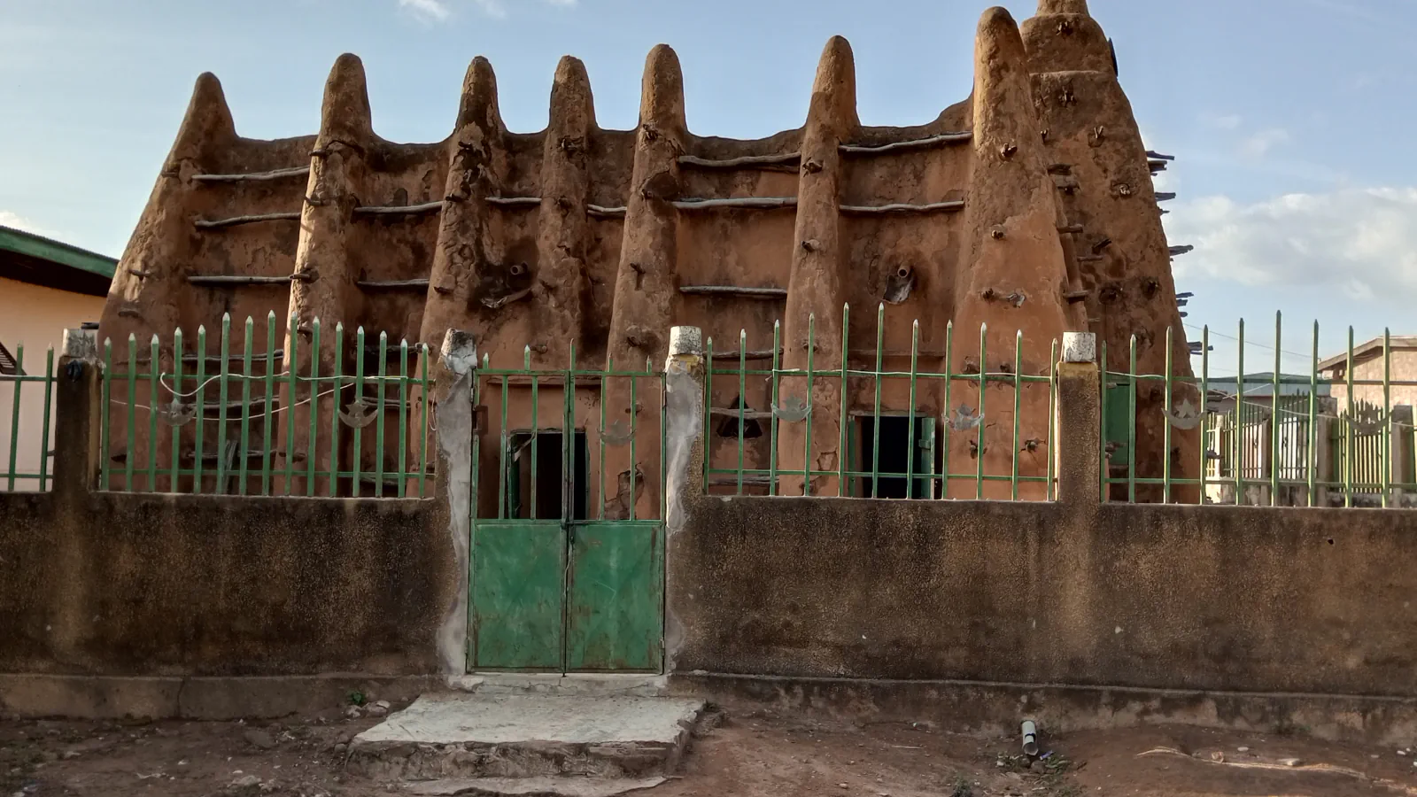 Earthen mosque with tall tapered buttresses and wooden beams projecting from the walls, behind a low wall and green metal gate.