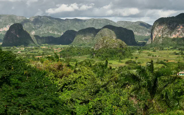Rounded, forested limestone mogotes rise above a green valley of fields, with low clouds over the distant hills.