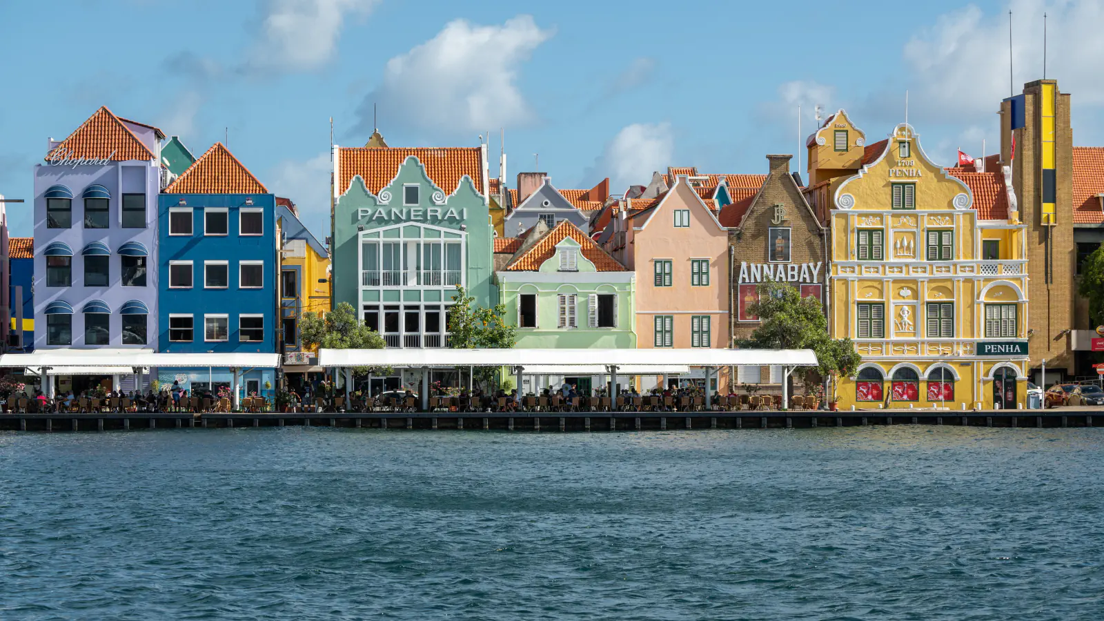 Waterfront row of tall gabled buildings painted lilac, blue, green, pink and yellow along the Handelskade in Punda, Willemstad, seen across the harbour water