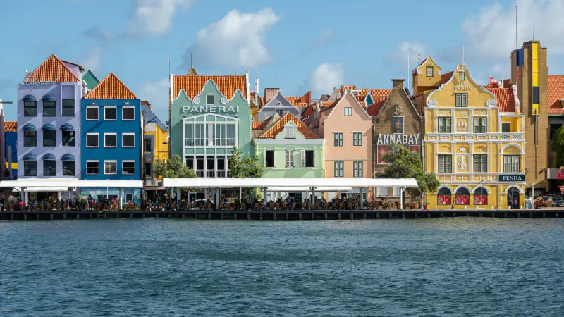 Waterfront row of tall gabled buildings painted lilac, blue, green, pink and yellow along the Handelskade in Punda, Willemstad, seen across the harbour water