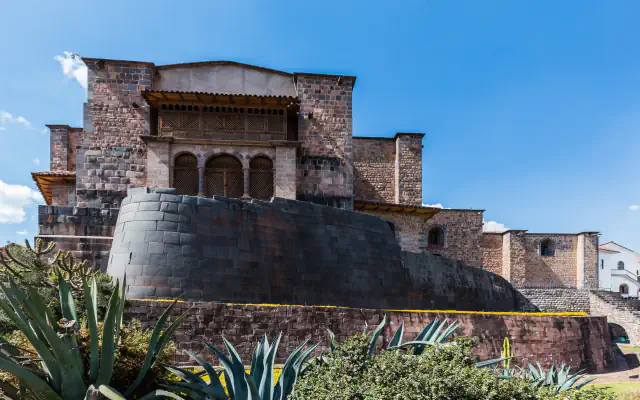 Dark, tightly fitted curved Inca stone wall beneath the brown stone walls and wooden balcony of the Santo Domingo convent, under a blue sky