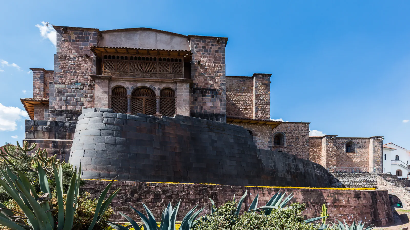 Dark, tightly fitted curved Inca stone wall beneath the brown stone walls and wooden balcony of the Santo Domingo convent, under a blue sky