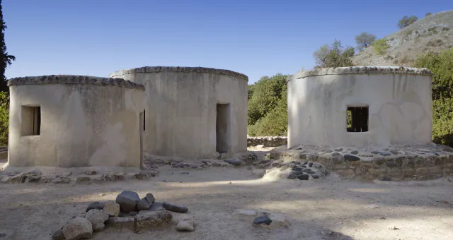 Three round, plastered buildings with flat tops and small windows stand among low stone walls under a blue sky, with a dry hillside behind