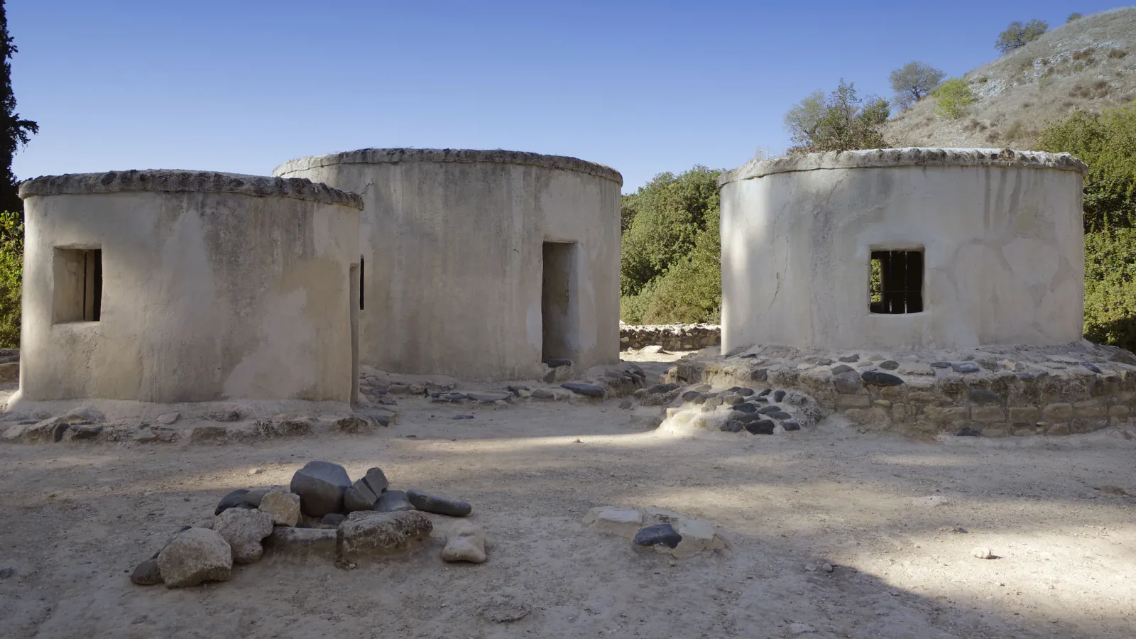 Three round, plastered buildings with flat tops and small windows stand among low stone walls under a blue sky, with a dry hillside behind