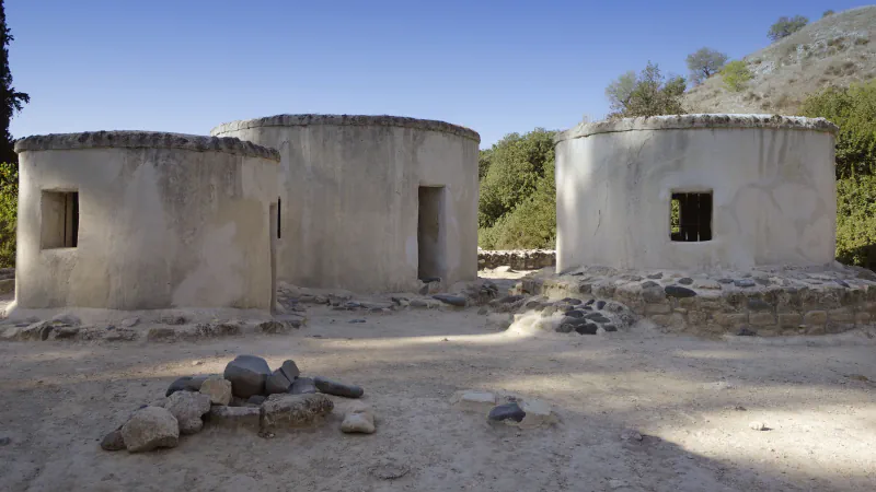 Three round, plastered buildings with flat tops and small windows stand among low stone walls under a blue sky, with a dry hillside behind