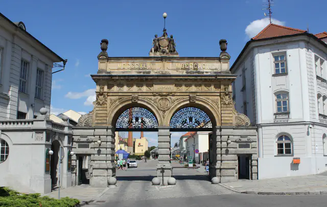 Stone gateway of the Plzeň brewery with two arches, topped by a sculpture group and inscribed with the Roman numerals for 1842 and 1892, with brewery buildings and a chimney beyond