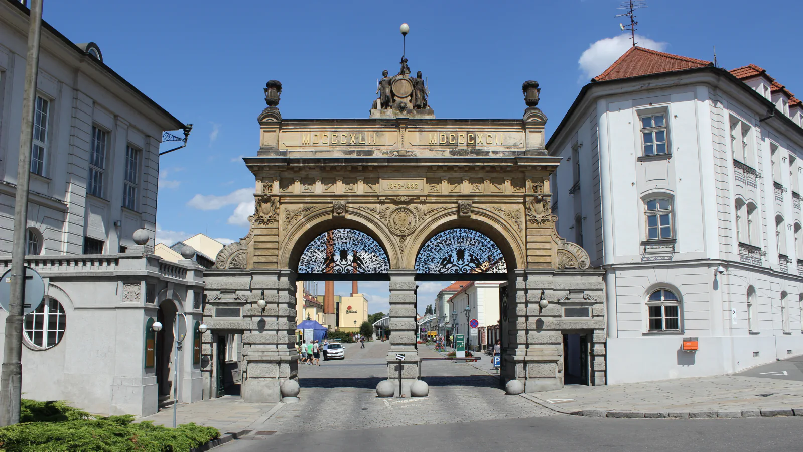 Stone gateway of the Plzeň brewery with two arches, topped by a sculpture group and inscribed with the Roman numerals for 1842 and 1892, with brewery buildings and a chimney beyond