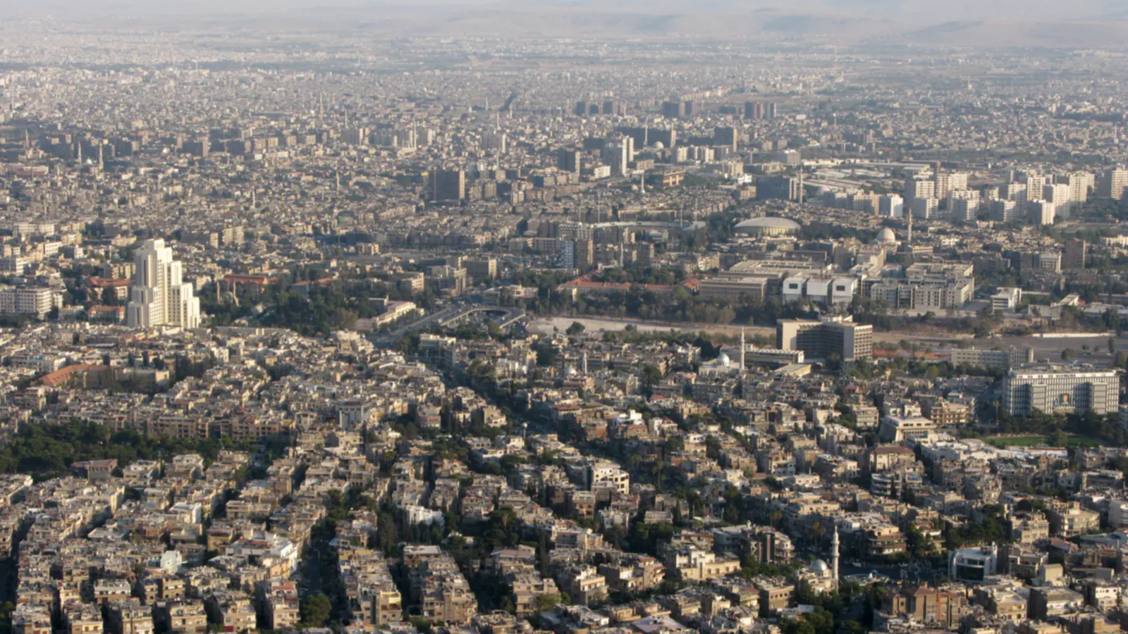 A hazy elevated view of Damascus, with dense blocks of buildings stretching to the horizon.