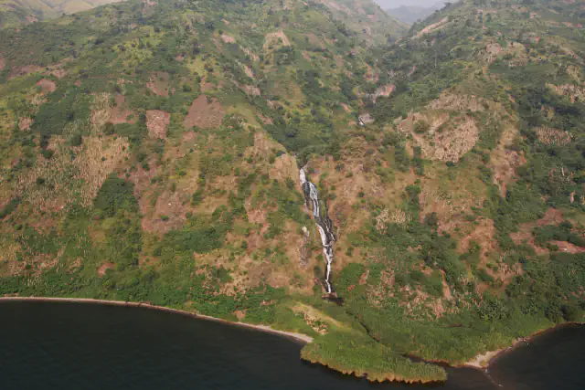 Aerial view of a green and brown mountainside dropping to a dark lake, with a narrow waterfall running down the slope and a green point jutting into the water.