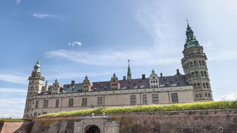 Sandstone wing of Kronborg Castle with towers and green copper spires rising above a grass-topped brick rampart and gateway