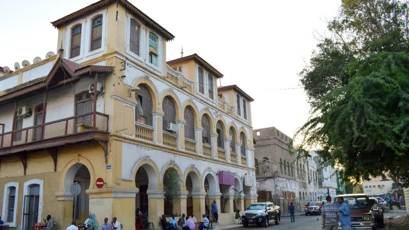 A corner building on Place Ménélik in Djibouti City: three storeys of yellow and white arcades and shuttered arched windows above covered ground-floor galleries, with a timber balcony and a tree-lined street