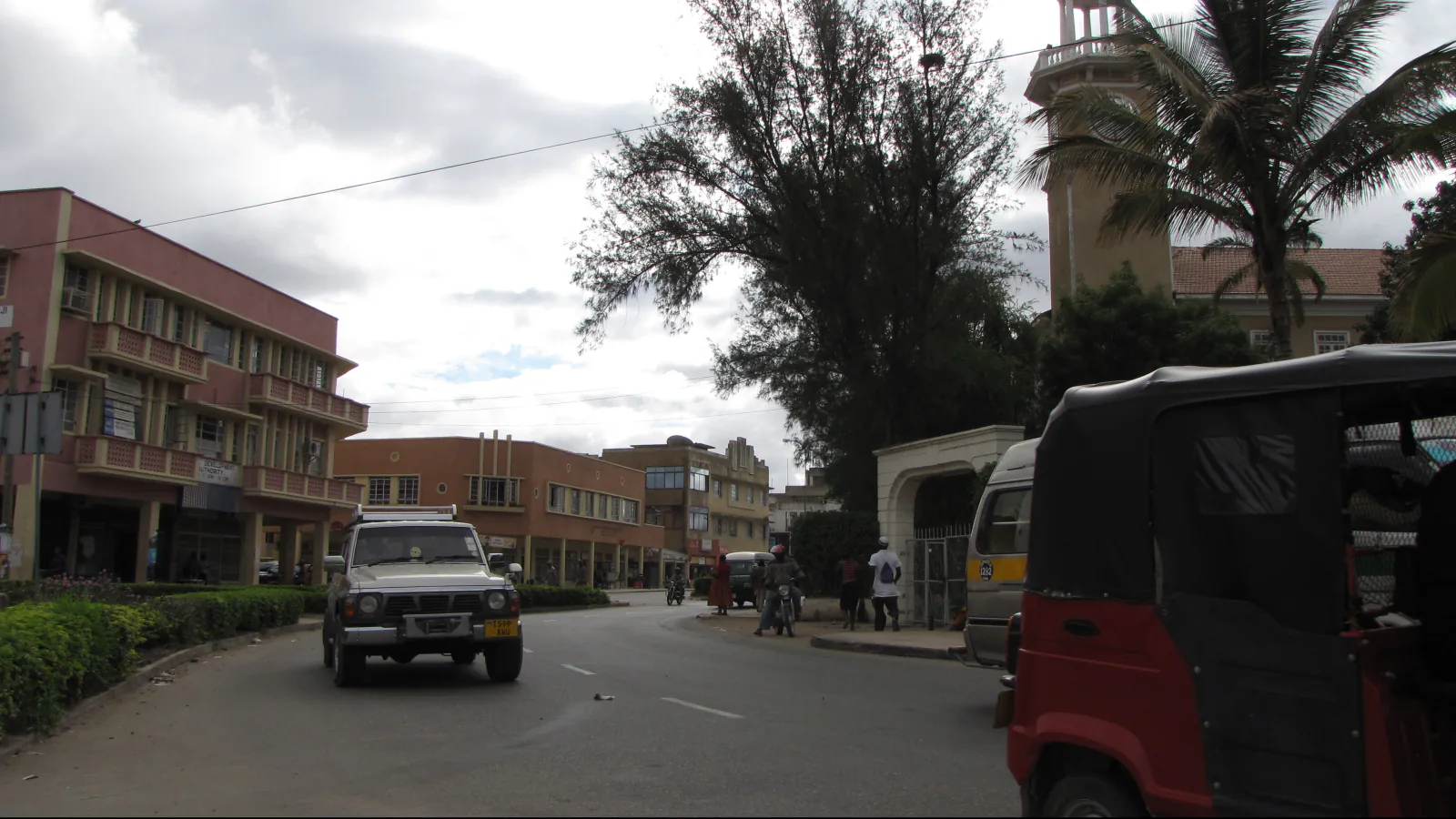 A street in Dodoma with low-rise buildings, trees, a tower at the right edge and vehicles under a cloudy sky.
