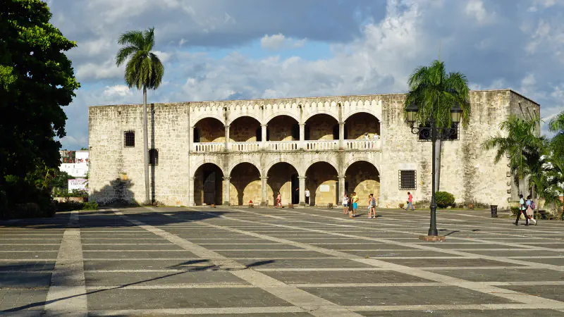The Alcázar de Colón across a paved plaza: a two-storey pale-stone palace with arcades on both floors; palms stand to the right and a tall palm to the left beneath a cloudy blue sky, with a few people crossing the plaza.