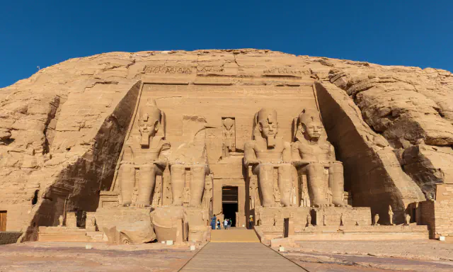 Four seated colossal statues of Ramesses II flank the doorway of the Great Temple at Abu Simbel, set against a rock face under a clear blue sky; one statue is broken above the waist