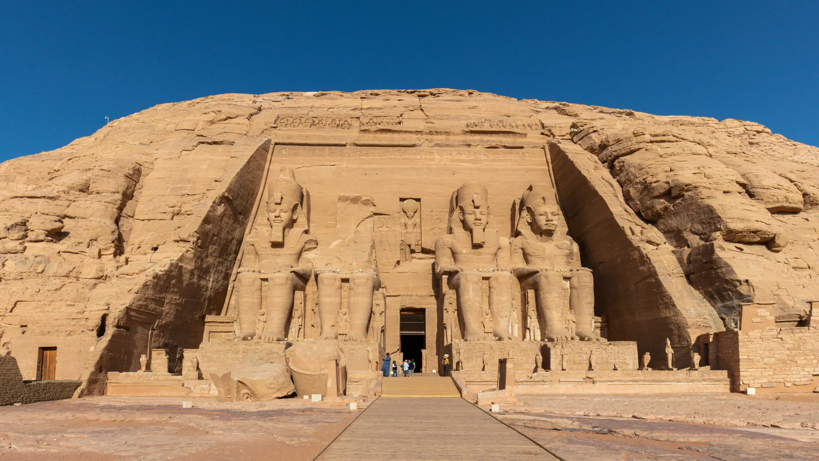 Four seated colossal statues of Ramesses II flank the doorway of the Great Temple at Abu Simbel, set against a rock face under a clear blue sky; one statue is broken above the waist