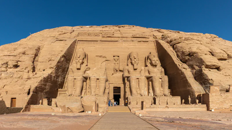 Four seated colossal statues of Ramesses II flank the doorway of the Great Temple at Abu Simbel, set against a rock face under a clear blue sky; one statue is broken above the waist