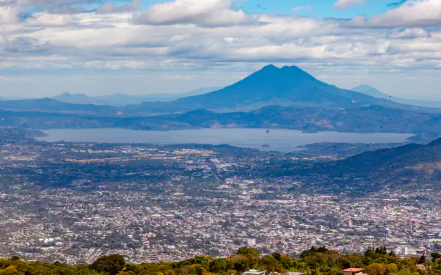 Wide view across the city of San Salvador to a lake in a broad basin, with a twin-peaked volcano rising behind it under a cloudy blue sky