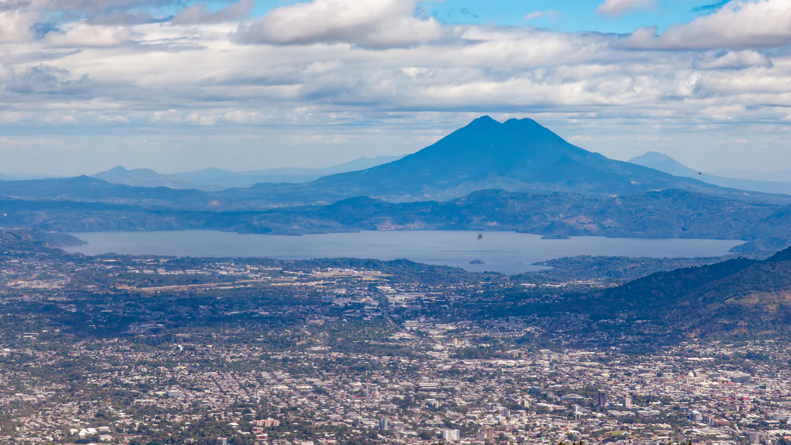 Wide view across the city of San Salvador to a lake in a broad basin, with a twin-peaked volcano rising behind it under a cloudy blue sky