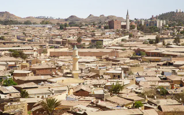 Rooftops of Asmara in clear light: a pale minaret with a green cap rises among low corrugated-iron and brick roofs, with a church spire, taller buildings and brown hills behind