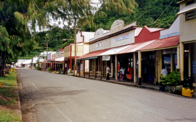 A quiet street of weatherboard shopfronts with red corrugated-iron roofs in Levuka, with a feathery-leaved tree at the left and forested hills behind