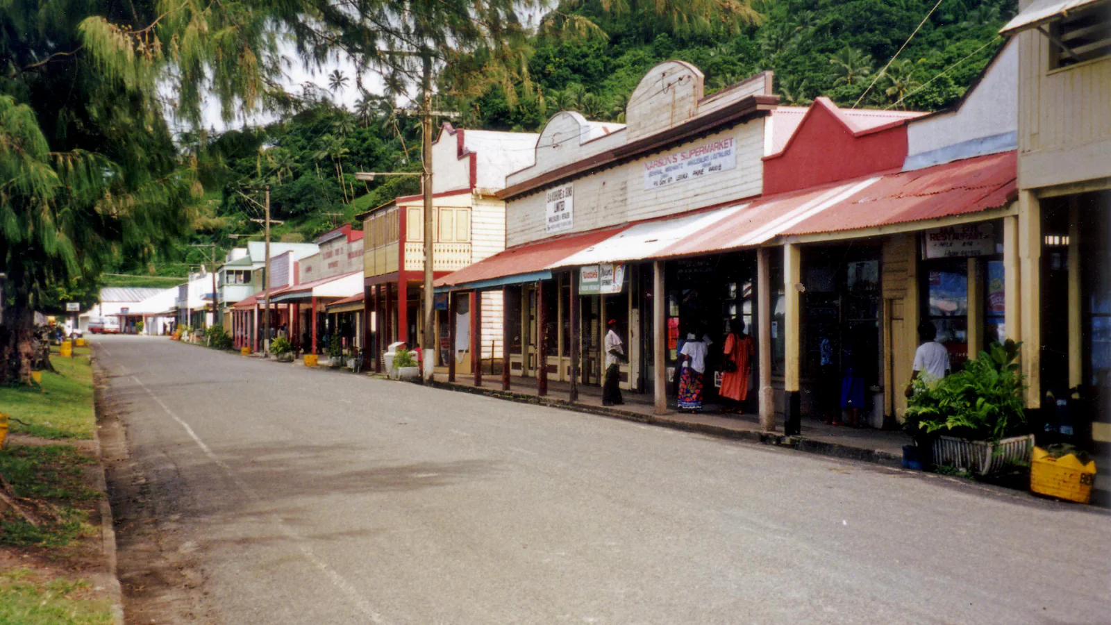 A quiet street of weatherboard shopfronts with red corrugated-iron roofs in Levuka, with a feathery-leaved tree at the left and forested hills behind