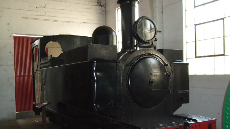 A black steam locomotive with a large front lamp and a red buffer beam inside a whitewashed museum shed.
