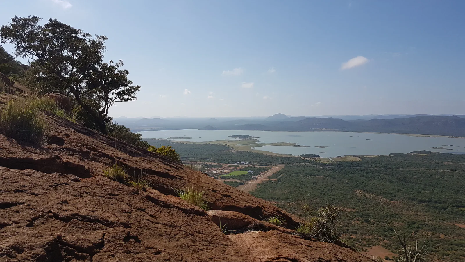 Looking down from a reddish granite slope with a tree and grass tufts on the left, over bushland to the Gaborone Dam reservoir; low hills line the horizon.