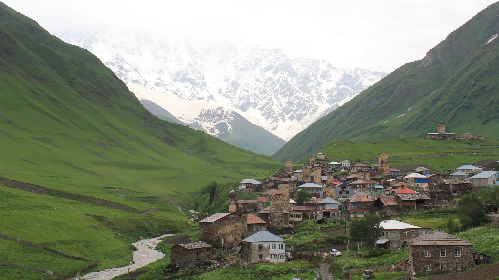 Svan tower houses and village buildings at Ushguli above a stream, with snow-covered peaks behind