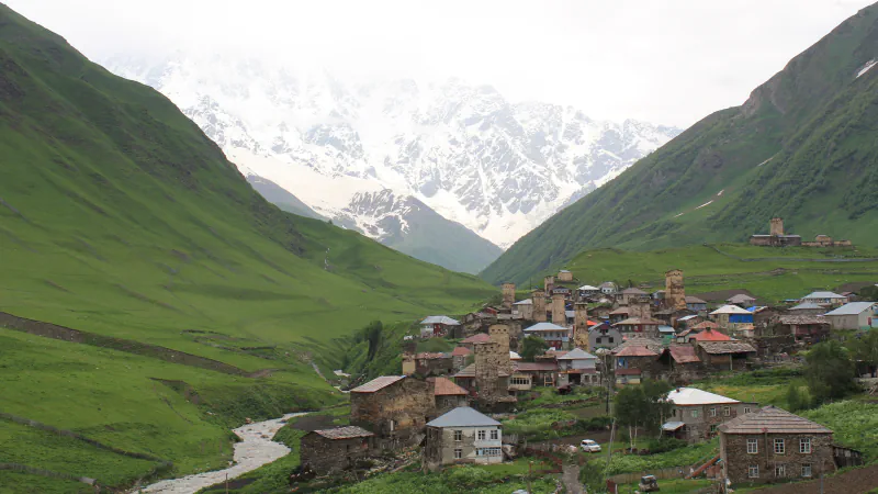 Svan tower houses and village buildings at Ushguli above a stream, with snow-covered peaks behind