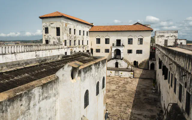 View down into Elmina Castle courtyard, bounded by whitewashed walls. A red-tiled building stands at the far end; the wall at right has a continuous top with repeated narrow openings.