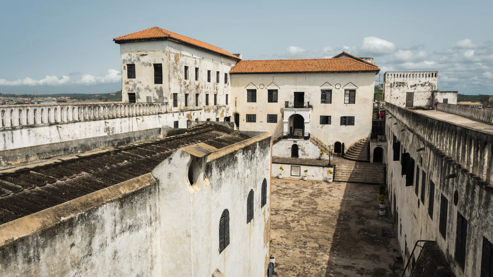 View down into Elmina Castle courtyard, bounded by whitewashed walls. A red-tiled building stands at the far end; the wall at right has a continuous top with repeated narrow openings.