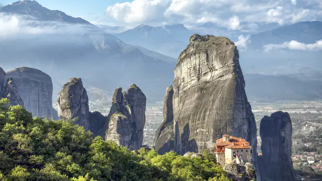 Sandstone pillars at Meteora rising above green forest, with a red-roofed monastery on a rock ledge at lower right and mountains beyond