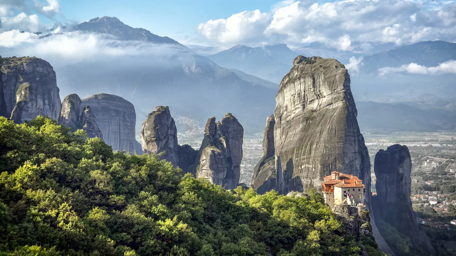 Sandstone pillars at Meteora rising above green forest, with a red-roofed monastery on a rock ledge at lower right and mountains beyond