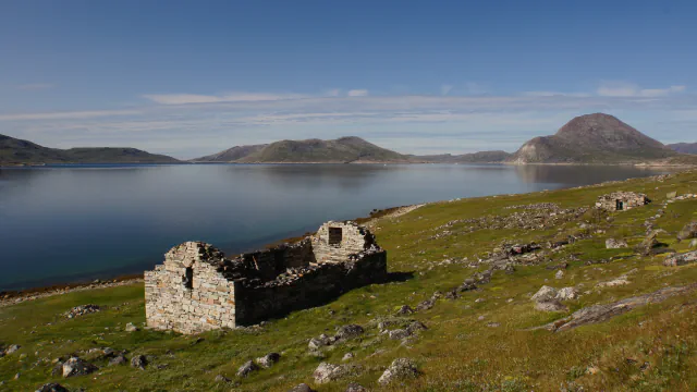Roofless stone ruin on a grassy shore beside a calm fjord, with low mountains behind