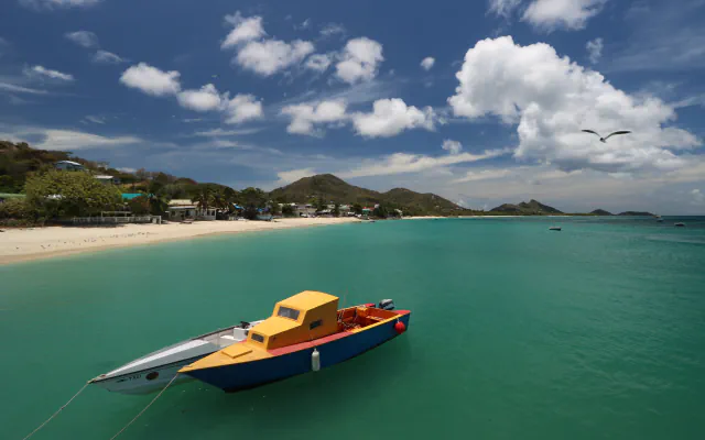 A yellow-cabin boat with a red deck and dark blue hull floats beside a smaller white craft marked TAXI; sandy shore, houses, green hills and clouds fill the background